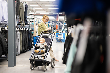 Image showing Casualy dressed mother choosing sporty shoes and clothes products in sports department of supermarket store with her infant baby boy child in stroller.