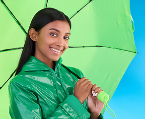 Image showing Portrait, smile and female with an umbrella in a studio with a stylish, trendy and fashion green rain coat. Life insurance, face and Indian woman model with winter outfit isolated by blue background.