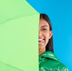 Image showing Umbrella, silly and woman portrait with tongue out in studio with raincoat and happiness. Fun, face and playful young female in isolated blue background with comedy, emoji and funny joke with a smile