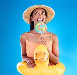 Image showing Pool float, happy woman and lollipop lick in a studio with sweet, swimsuit and sweets. Isolated, blue background and holiday of a young female with happiness, licking candy and feeling funny or silly