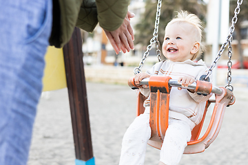 Image showing Mother pushing her cheerful infant baby boy child on a swing on sandy beach playground outdoors on nice sunny cold winter day in Malaga, Spain.