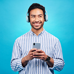 Image showing Headphones, music and thinking man with phone in studio for streaming, audio and subscription against blue background. Radio, smile and happy asian male online for podcast, track and internet app
