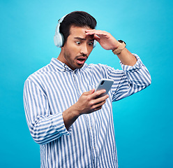 Image showing Phone, headphones and shock with a man on a blue background in studio reading news on social media. Wow, surprise or podcast with a male listening to audio in disbelief at hearing mind blowing gossip