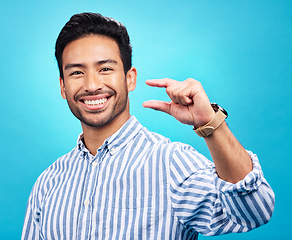 Image showing Small, hand and gesture of a man portrait with a smile in studio showing a tiny measurement. Isolated, blue background and Asian model with happiness, positive opinion and hands show of pinch size