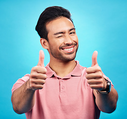 Image showing Portrait, thumbs up or wink by man in studio in positive, feedback or review on blue background. Face, smile and hand gesture by Asian male model showing yes, agreement or vote emoji while isolated