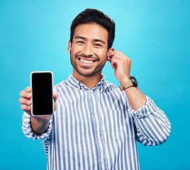 Image showing Asian man, phone and mockup screen with earphones for music listening, call or audio against a blue studio background. Portrait of happy male showing smartphone display for sound app or communication