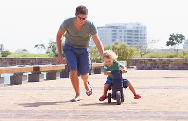 Image showing Happy, running and motorbike with child and father playing in park for bonding, laugh and helping. Smile, happiness and energy with man and young boy riding on scooter for playful, learning or summer