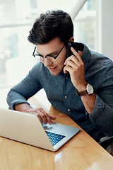 Image showing Let me check that for you. Cropped shot of a handsome young businessman sitting and using his cellphone while working on his laptop in the office.