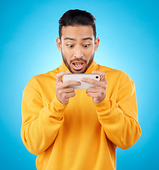 Image showing Phone, reading and a shocked man in studio with a secret, gossip or fake news on social media. Male asian model watch video or post on smartphone for wow, surprise or chat on a blue background