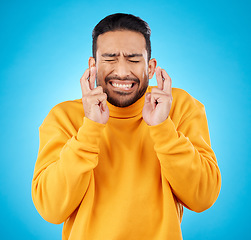 Image showing Nervous, eyes closed and fingers crossed by asian man with anxiety in studio for news, feedback or review on blue background. Hand, emoji and guy with stress for results, competition or giveaway