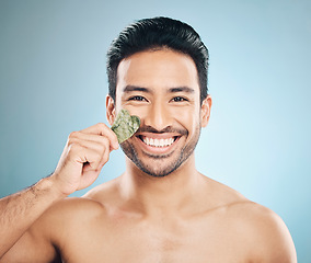 Image showing Face, man and beauty, gua sha and cosmetic tools, smile in portrait and natural treatment on blue background. Skincare, healthy glow and jade stone, dermatology and wellness with male model in studio