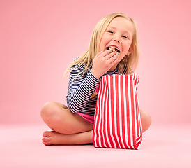 Image showing Popcorn eating, portrait and happy girl in a studio with pink background sitting with movie snacks. Food taste, happiness and hungry young child with a paper bag and chips feeling relax with a smile