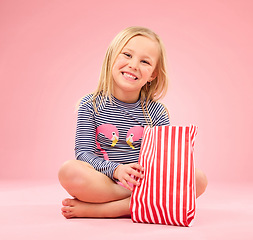 Image showing Popcorn, food and happy girl portrait in a studio with pink background sitting with movie snacks. Snack, happiness and hungry child with a paper bag and chips eating and feeling relax with a smile