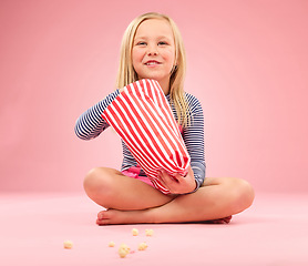 Image showing Popcorn, snack and happy girl in a studio with pink background sitting with movie snacks. Food, happiness and hungry young child with a paper bag and chips eating and feeling relax with a smile