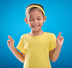 Image showing Child, hand and fingers crossed in studio for hope, wish or good luck against a blue background. Girl, eyes closed and emoji hands, praying and wishing, happy and smile while posing on isolated space