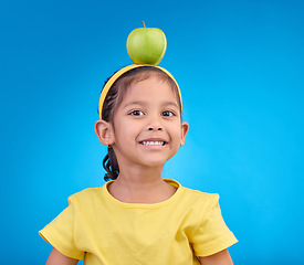 Image showing Portrait, smile and girl with apple on head on blue background for nutrition, healthy eating and diet. Food, youth and face of happy young girl in studio with fruit for organic, vitamins and wellness