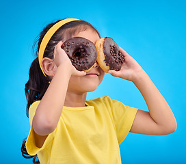 Image showing Doughnuts, eyes and face of playful child with food isolated against a studio blue background with a smile. Adorable, happy and cute young girl or kid excited for sweet sugar donut calories