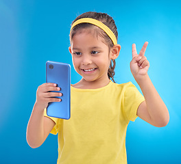 Image showing Phone, girl kid and peace sign selfie in studio isolated on a blue background. Technology, v hand emoji and smile of child with mobile smartphone to take pictures for happy memory or social media.