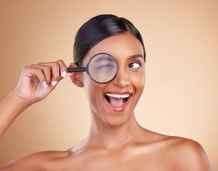 Image showing Face, beauty or magnifying glass with a model woman in studio on beige background searching for skincare. Facial, eye wink or excited Indian girl looking for luxury self care cosmetics or makeup