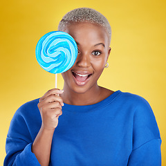 Image showing Excited, portrait and black woman with a lollipop in studio for dessert, sweet treat or craving. Happy, surprise and face of African female model posing with blue candy isolated by yellow background.