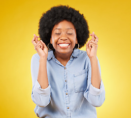 Image showing Happy, smile and fingers crossed by black woman in studio for wish, hope and good luck against yellow background. Eyes closed, hand and emoji by excited female waiting, optimistic and hopeful gesture