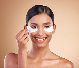 Image showing Eye patch, beauty and portrait of a woman in studio for skincare, dermatology and cosmetics. Happy Indian female model with smile and collagen gel mask for self care facial on brown background