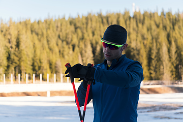 Image showing Handsome male athlete with cross country skis, taking fresh breath and having break after hard workout training in a snowy forest. Checking smartwatch. Healthy winter lifestyle.