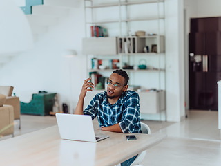 Image showing African American man in glasses sitting at a table in a modern living room, using a laptop for business video chat, conversation with friends and entertainment