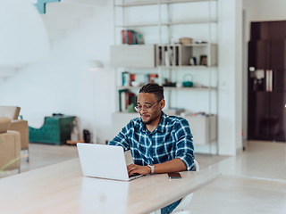 Image showing African American man in glasses sitting at a table in a modern living room, using a laptop for business video chat, conversation with friends and entertainment