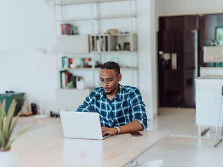 Image showing African American man in glasses sitting at a table in a modern living room, using a laptop for business video chat, conversation with friends and entertainment