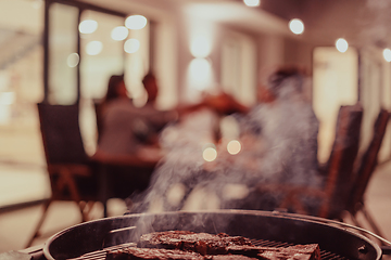 Image showing Close-up photo of delicious meat being grilled. In the background, friends and family are sitting and waiting for a meal