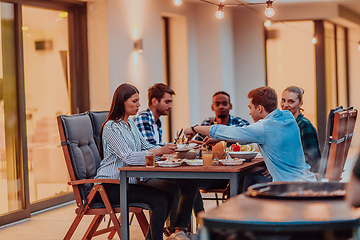 Image showing A group of young diverse people having dinner on the terrace of a modern house in the evening. Fun for friends and family. Celebration of holidays, weddings with barbecue.