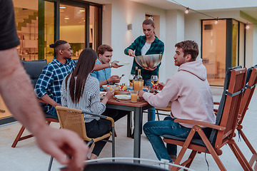 Image showing A group of young diverse people having dinner on the terrace of a modern house in the evening. Fun for friends and family. Celebration of holidays, weddings with barbecue.