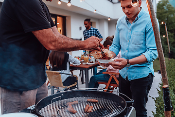 Image showing A group of friends and family barbecue together in the evening on the terrace in front of a large modern house