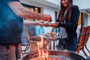 Image showing A group of friends and family barbecue together in the evening on the terrace in front of a large modern house