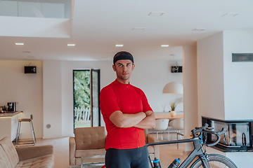 Image showing Photo of a determined triathlete standing in a modern large living room, behind him is his training bike