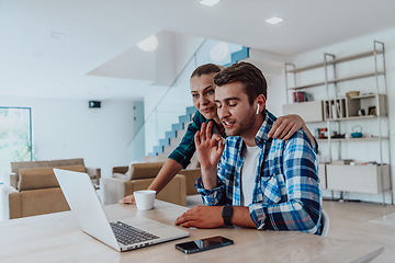 Image showing A young married couple is talking to parents, family and friends on a video call via a laptop while sitting in the living room of their modern house