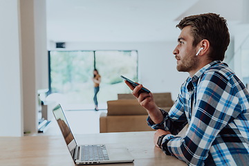 Image showing The man sitting at a table in a modern living room, using a smartphone and laptop for business video chat, conversation with friends and entertainment