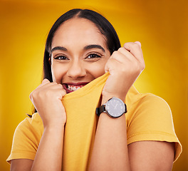 Image showing Portrait, shy and playful with a woman on a yellow background in studio pulling on her t-shirt. Face, fashion or funny and an attractive young female covering her mouth with trendy clothes for style