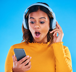 Image showing Headphones, cellphone and shocked woman in a studio listening to a podcast, music or radio. Surprise, technology and female model streaming an album, song or playlist with a phone by blue background.