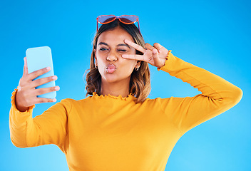 Image showing Selfie, studio and peace sign of a woman influencer taking a profile picture for social media. Isolated, blue background and silly hands gesture of a gen z female with a kiss feeling happy and fun