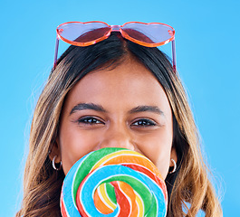 Image showing Portrait, giant lollipop and a woman on a blue background in studio wearing heart glasses for fashion. Face, candy and sweet with an attractive young female eating a snack while feeling happy