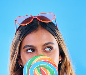 Image showing Thinking, lollipop and a woman on a blue background in studio wearing heart glasses for fashion. Idea, candy and sweet with an attractive young female eating a giant snack while feeling thoughtful