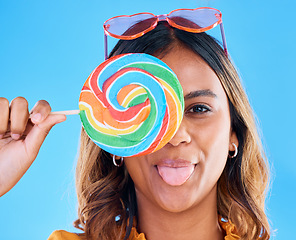 Image showing Portrait, lollipop and tongue with a woman on a blue background in studio wearing heart glasses for fashion. Funny face, candy and sweet with a young female eating a giant snack while feeling silly