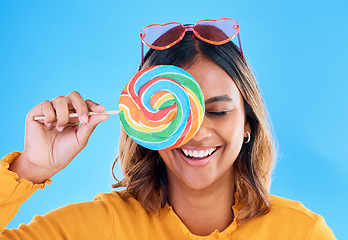 Image showing Laughing, lollipop and a woman on a blue background in studio wearing heart glasses for fashion. Funny, candy and sweet with an attractive young female eating a giant snack while feeling silly