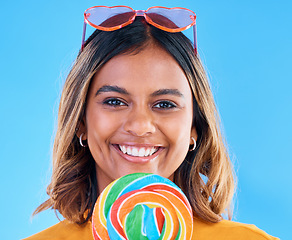 Image showing Portrait, lollipop and a woman smiling on a blue background in studio wearing heart glasses for fashion. Face, candy and sweet with a happy young female eating a giant snack while feeling positive