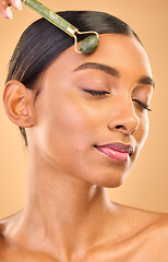 Image showing Face, skincare roller and woman with eyes closed in studio isolated on a brown background. Dermatology, facial massage and Indian female model with jade crystal for healthy skin treatment and beauty.