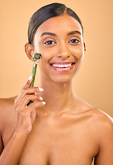 Image showing Face portrait, skincare roller and woman in studio isolated on a brown background. Dermatology, facial massage and happy Indian female model with jade crystal for healthy skin treatment and beauty.