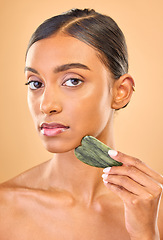 Image showing Face portrait, skincare and woman with gua sha in studio isolated on a brown background. Dermatology, massage and serious Indian female model with jade crystal or stone for healthy skin treatment.