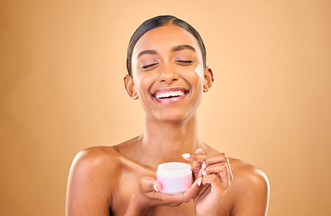 Image showing Face, skincare and woman with cream jar in studio isolated on a brown background. Dermatology cosmetics, funny and happy Indian female apply lotion, creme and moisturizer product for healthy skin.
